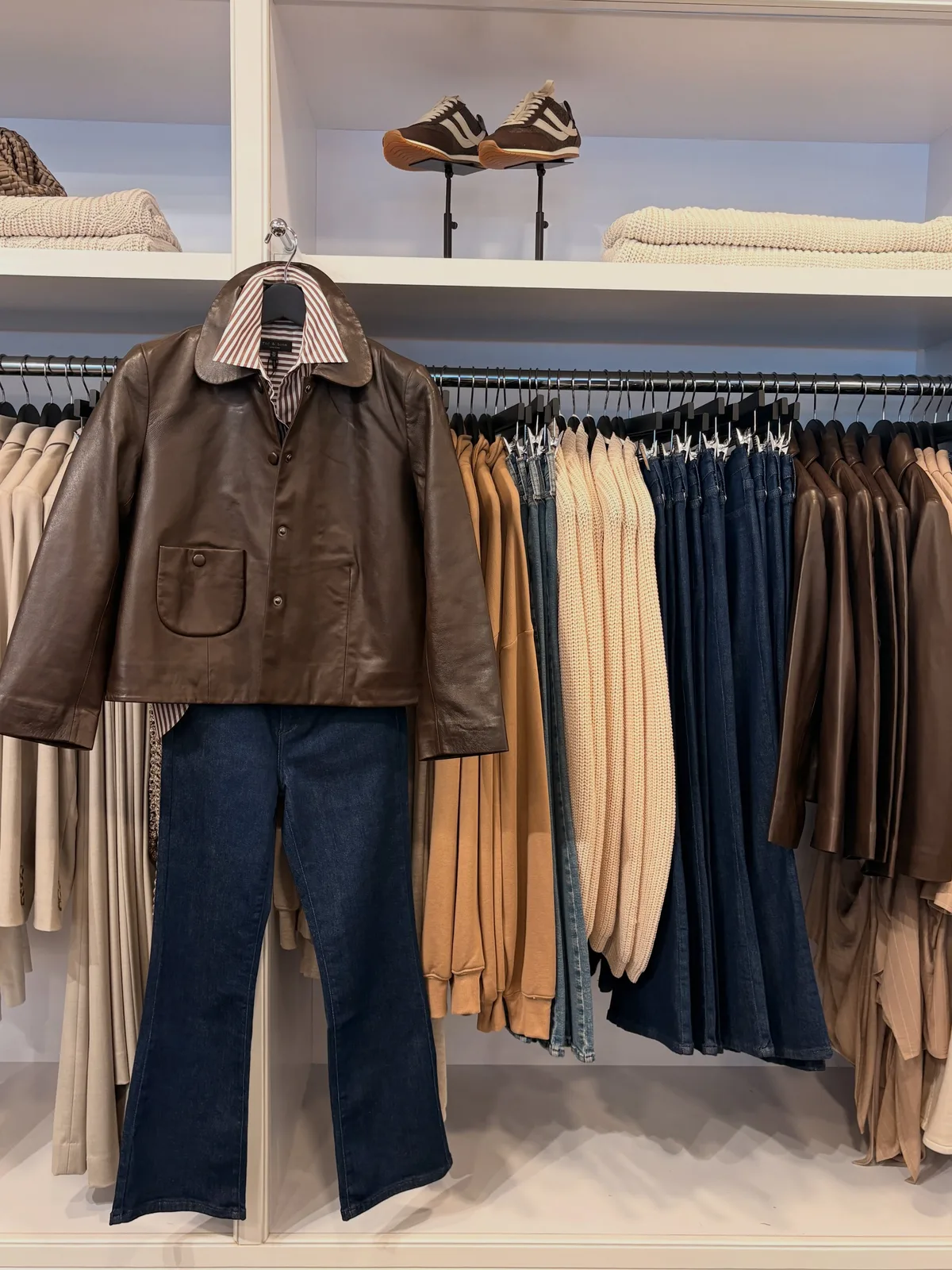 A brown leather jacket styled with dark jeans in front of a rack of knits and trousers